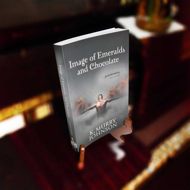 The novel Image of Emeralds and Chocolate by African American author K. Murry Johnson stands upright on a small wooden book stand. The cover shows a dramatic image of a man with outstretched arms surrounded by swirling color. The book is placed on a dark, polished table with a soft, blurred background.