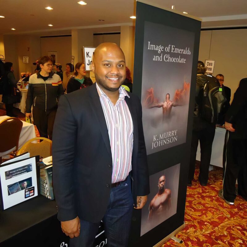 K. Murry Johnson stands smiling at a book signing event beside a display for his novel Image of Emeralds and Chocolate, surrounded by attendees in a convention-style setting.
