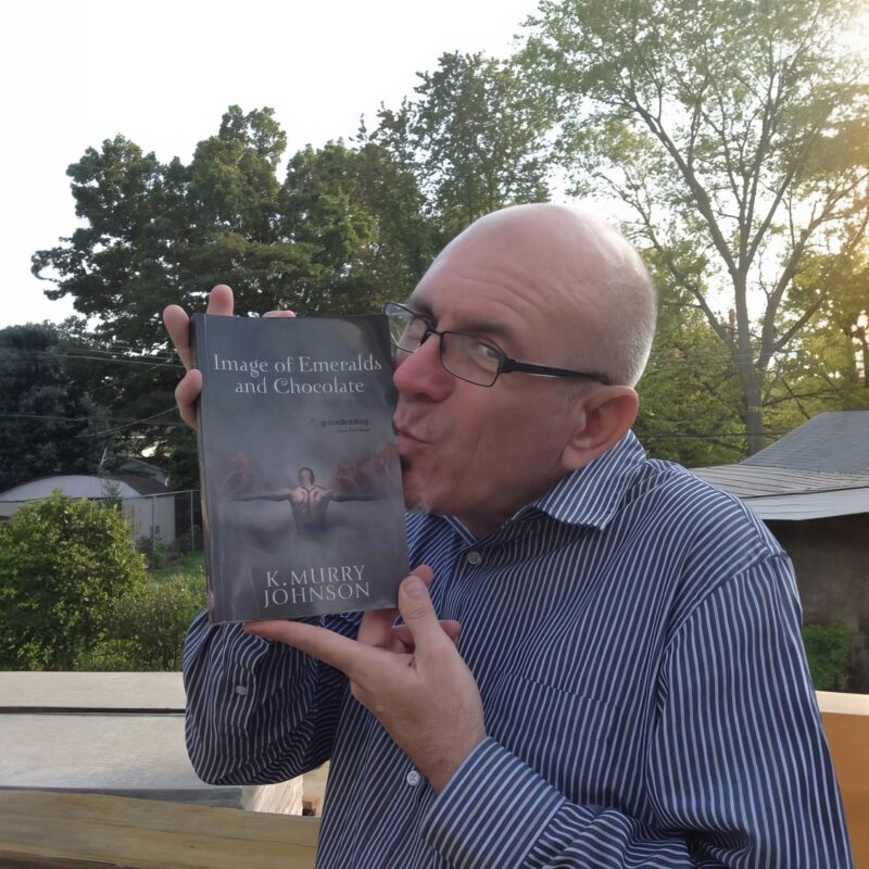 A reader holds and kisses a copy of Image of Emeralds and Chocolate by K. Murry Johnson while standing outdoors with trees in the background.