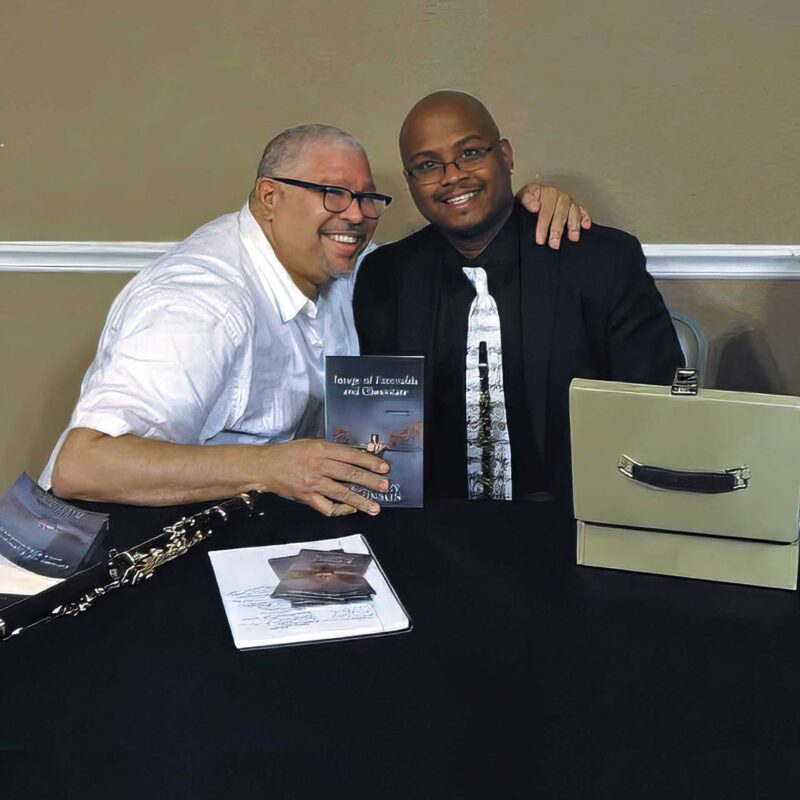 A reader sits beside K. Murry Johnson at a book signing table, holding a copy of Image of Emeralds and Chocolate, with books and a clarinet visible on the table.