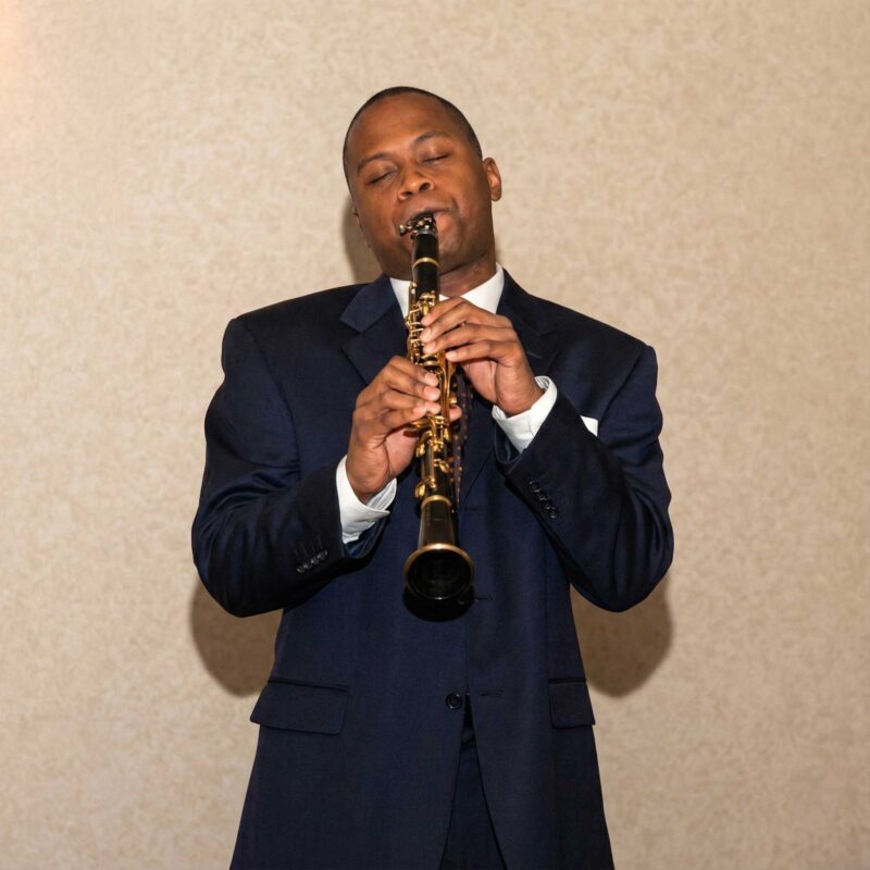 K. Murry Johnson plays the saxophone with eyes closed, wearing a dark suit against a neutral background, captured in an intimate moment of musical expression.